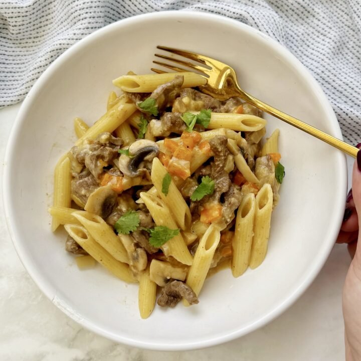 A hearty plate of cheesesteak pasta featuring thin slices of seared beef, colorful sautéed peppers, mushrooms, and onions, all tossed with whole wheat pasta in a creamy, cheesy sauce.