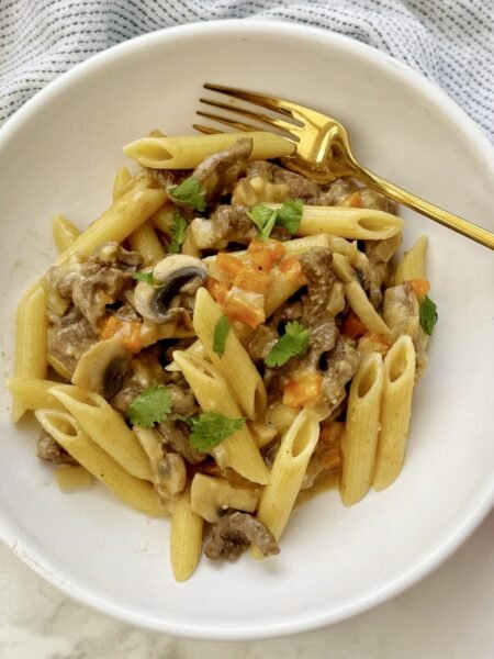 A hearty plate of cheesesteak pasta featuring thin slices of seared beef, colorful sautéed peppers, mushrooms, and onions, all tossed with whole wheat pasta in a creamy, cheesy sauce.