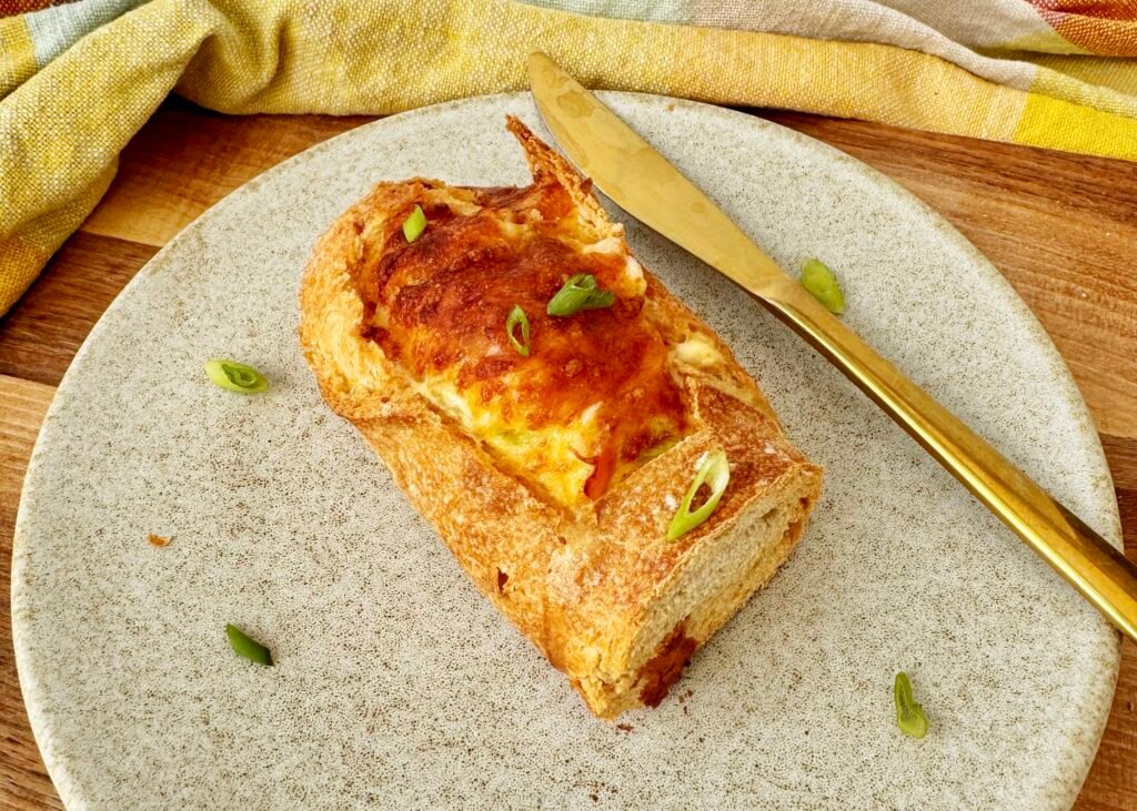 Cheesy Egg Baguette Bake sliced open to reveal a creamy egg filling with peppers and spring onions, topped with melted golden cheese, served on a rustic wooden board.
