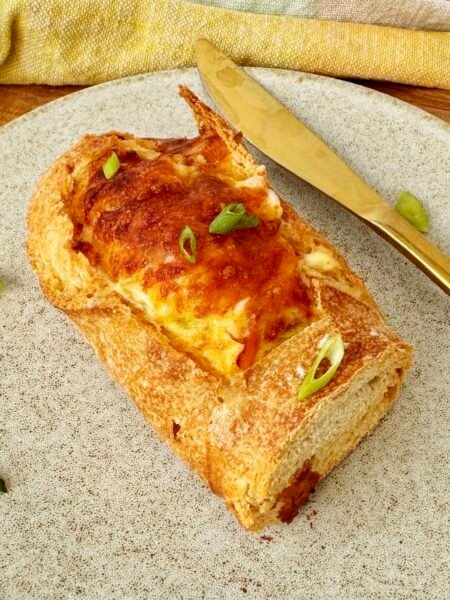 Cheesy Egg Baguette Bake sliced open to reveal a creamy egg filling with peppers and spring onions, topped with melted golden cheese, served on a rustic wooden board.