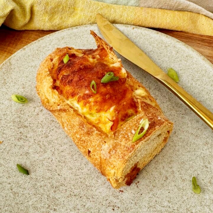 Cheesy Egg Baguette Bake sliced open to reveal a creamy egg filling with peppers and spring onions, topped with melted golden cheese, served on a rustic wooden board.