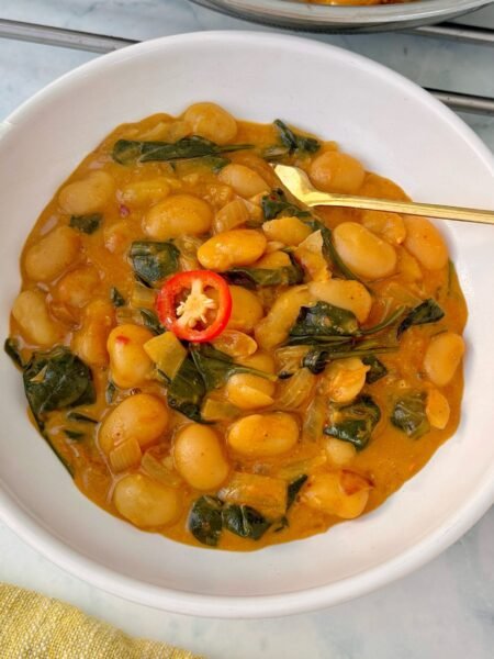 Bowl of creamy butter beans in coconut sauce garnished with spinach and herbs