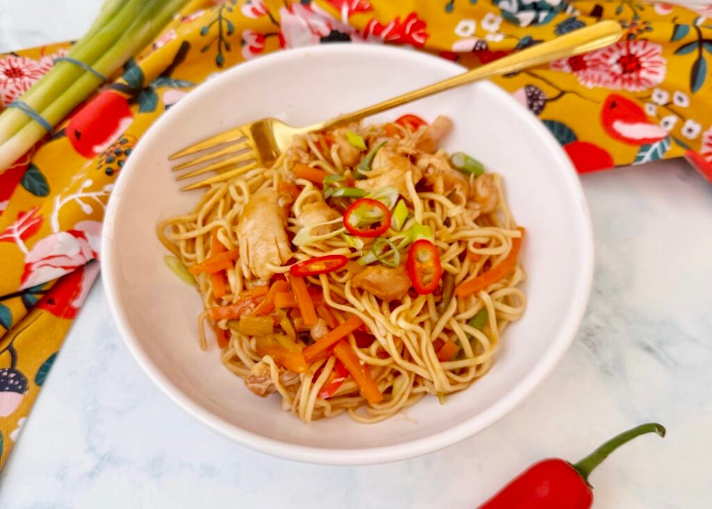 A bowl of Hot Honey Chicken Noodles with shredded chicken, egg noodles, julienned vegetables, and garnished with sesame seeds, chopped green onions, and red chilli slices, all coated in a glossy sweet and spicy sauce