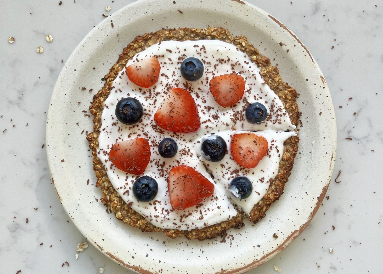 Healthy oatmeal breakfast pizza topped with yoghurt, strawberries, blueberries, and dark chocolate shavings.
