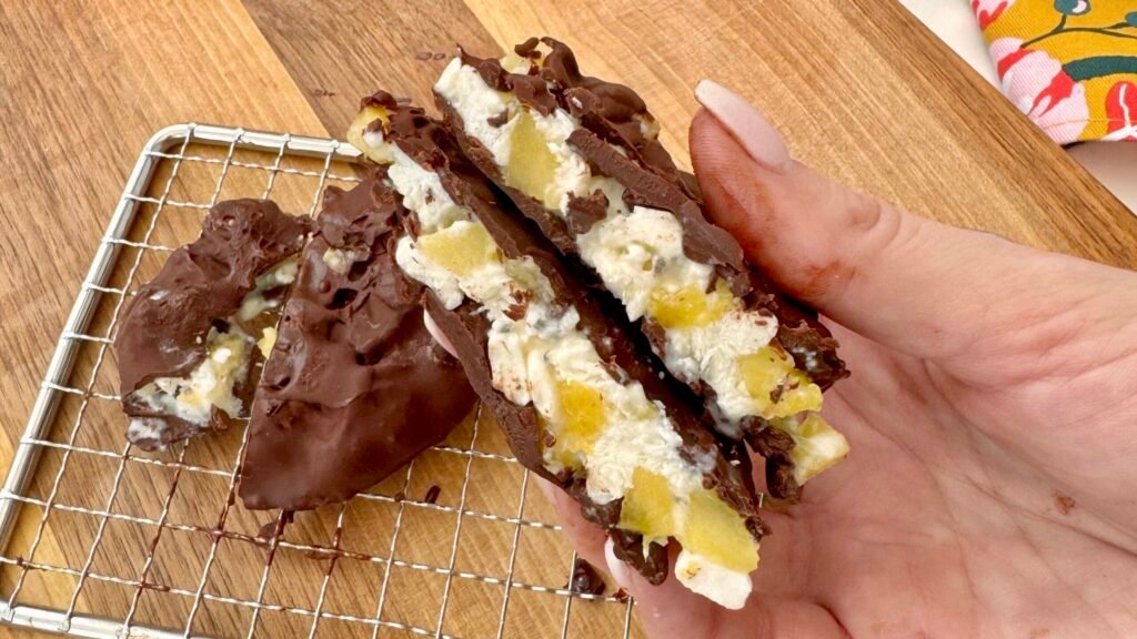 A close-up of four frozen pineapple and cottage cheese bites, coated in glossy dark chocolate, resting on parchment paper. The bites are round with visible pineapple chunks and sprinkled chia seeds, offering a contrast of textures and colors. Perfect as a healthy, high-protein snack or dessert.
