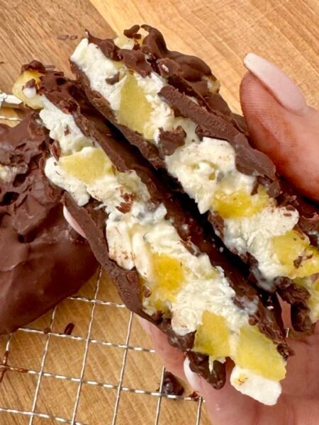 A close-up of four frozen pineapple and cottage cheese bites, coated in glossy dark chocolate, resting on parchment paper. The bites are round with visible pineapple chunks and sprinkled chia seeds, offering a contrast of textures and colors. Perfect as a healthy, high-protein snack or dessert.