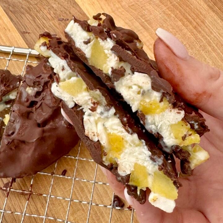A close-up of four frozen pineapple and cottage cheese bites, coated in glossy dark chocolate, resting on parchment paper. The bites are round with visible pineapple chunks and sprinkled chia seeds, offering a contrast of textures and colors. Perfect as a healthy, high-protein snack or dessert.