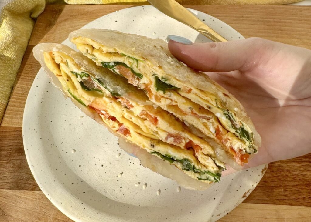 Golden brown rice paper omelette wrap sliced in half, filled with eggs, tomato, and spinach, garnished with sesame seeds.