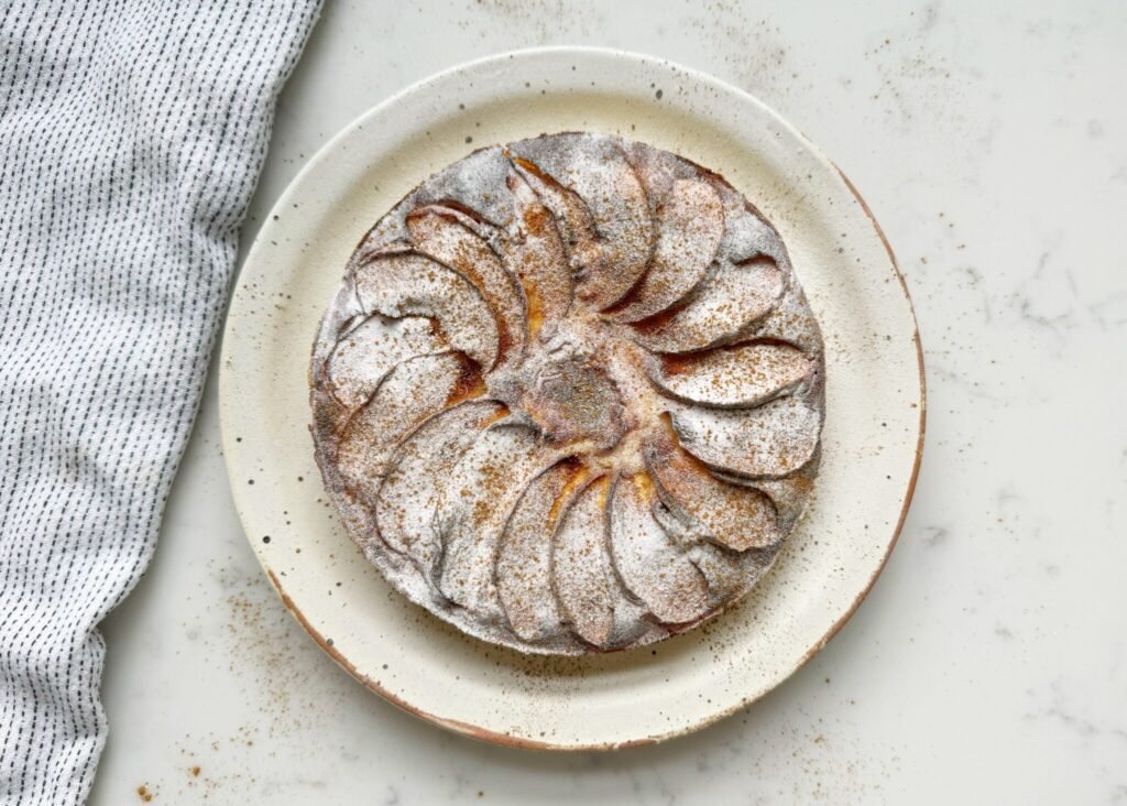 A golden-brown slice of spiced apple yoghurt cake topped with tender apple chunks and a light dusting of cinnamon. The cake looks moist and fluffy, served on a plate with a dollop of yoghurt and a sprinkle of nuts, creating a warm and wholesome presentation.