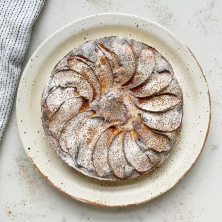 A golden-brown slice of spiced apple yoghurt cake topped with tender apple chunks and a light dusting of cinnamon. The cake looks moist and fluffy, served on a plate with a dollop of yoghurt and a sprinkle of nuts, creating a warm and wholesome presentation.