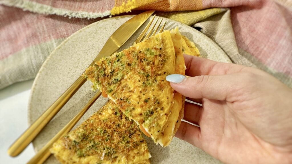 A golden omelette with crisp edges, filled with grated carrots, broccoli, and melted mozzarella, served hot on a white plate with a fresh herb garnish.