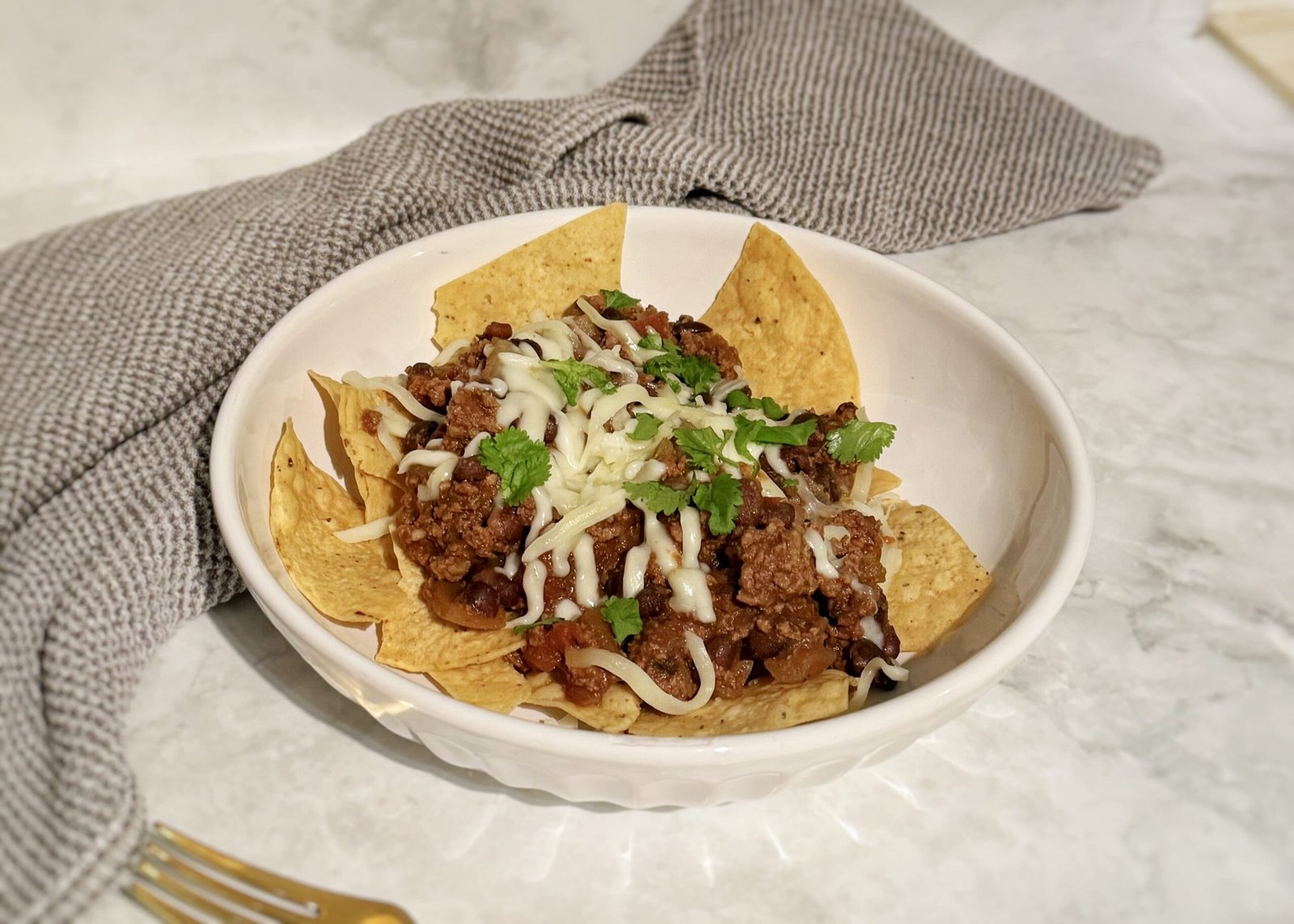 A hearty bowl of Slow Cooker Chilli Con Carne topped with melted cheese and fresh coriander, served with tortilla chips on the side.