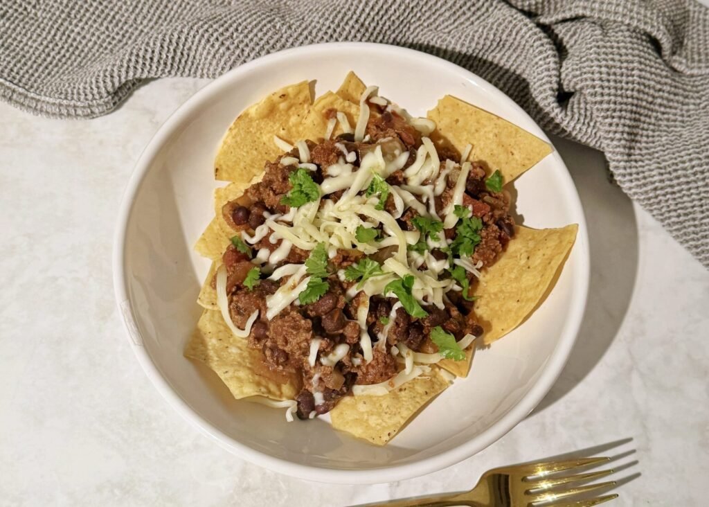 A hearty bowl of Slow Cooker Chilli Con Carne topped with melted cheese and fresh coriander, served with tortilla chips on the side.