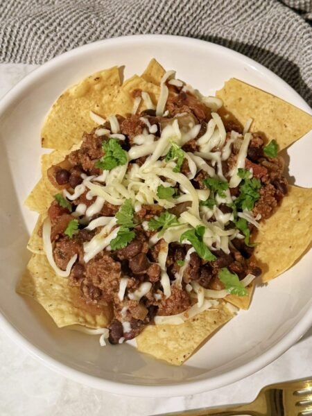 A hearty bowl of Slow Cooker Chilli Con Carne topped with melted cheese and fresh coriander, served with tortilla chips on the side.