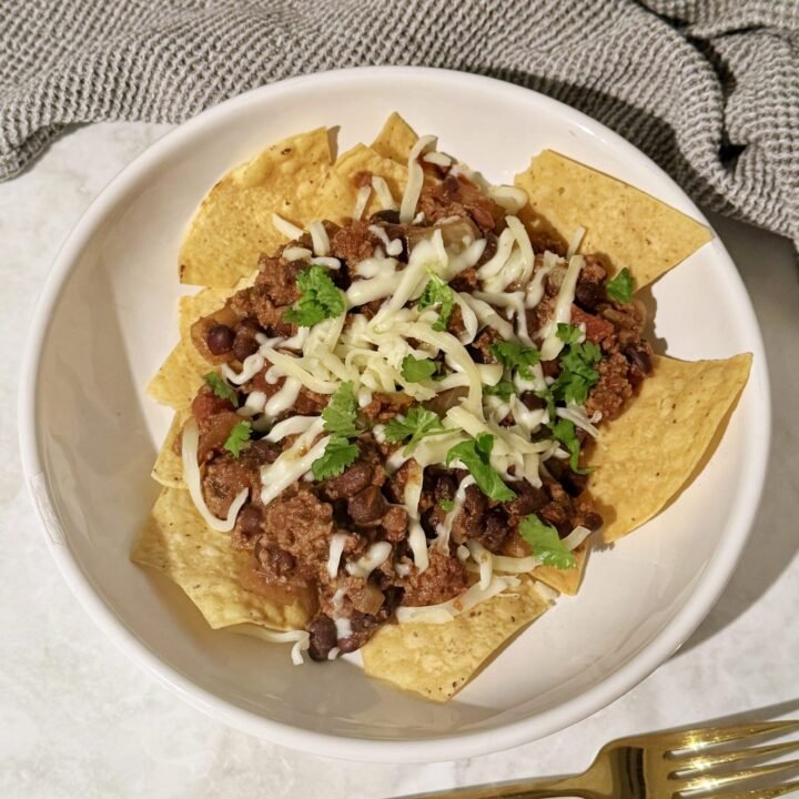 A hearty bowl of Slow Cooker Chilli Con Carne topped with melted cheese and fresh coriander, served with tortilla chips on the side.