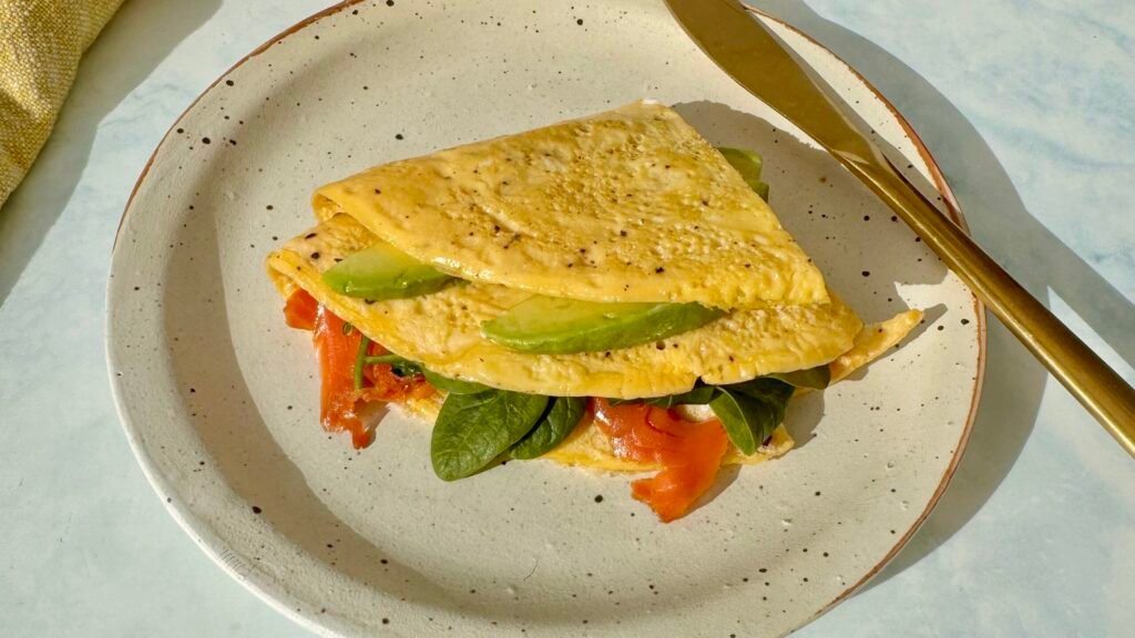 A golden egg wrap folded around layers of smoked salmon, avocado, spinach, and a drizzle of mayo, served on a plate.