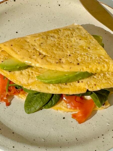 A golden egg wrap folded around layers of smoked salmon, avocado, spinach, and a drizzle of mayo, served on a plate.