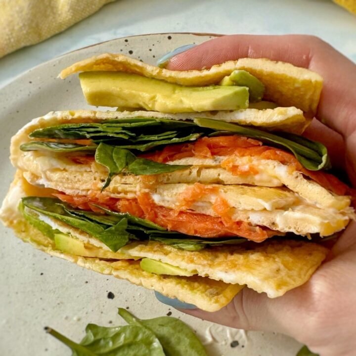 A golden egg wrap folded around layers of smoked salmon, avocado, spinach, and a drizzle of mayo, served on a plate.