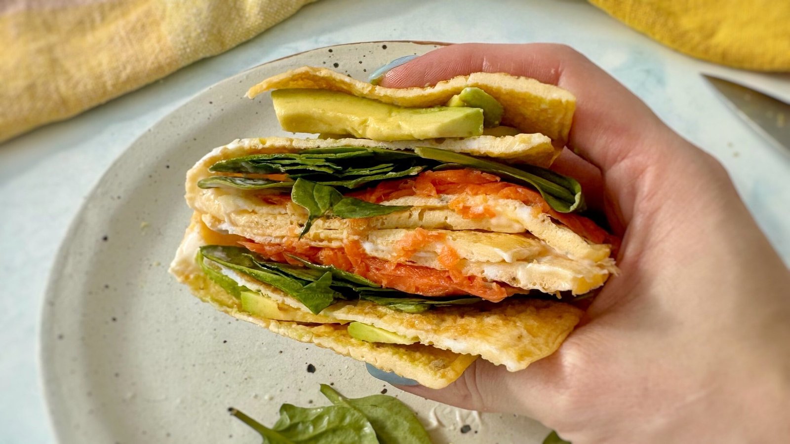 A golden egg wrap folded around layers of smoked salmon, avocado, spinach, and a drizzle of mayo, served on a plate.