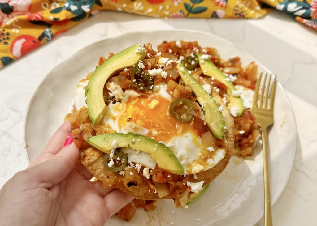 A plate of Huevos Rancheros with a crispy tortilla base, runny fried egg, refried beans, tomato salsa, avocado slices, and crumbled feta, garnished with coriander and lime wedges.