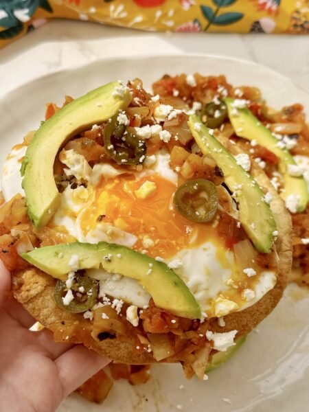 A plate of Huevos Rancheros with a crispy tortilla base, runny fried egg, refried beans, tomato salsa, avocado slices, and crumbled feta, garnished with coriander and lime wedges.