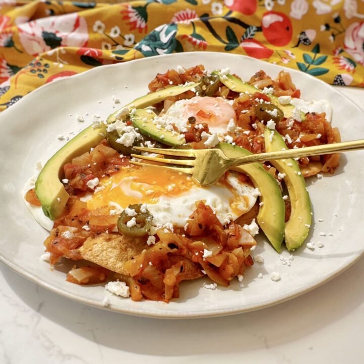 A plate of Huevos Rancheros with a crispy tortilla base, runny fried egg, refried beans, tomato salsa, avocado slices, and crumbled feta, garnished with coriander and lime wedges.