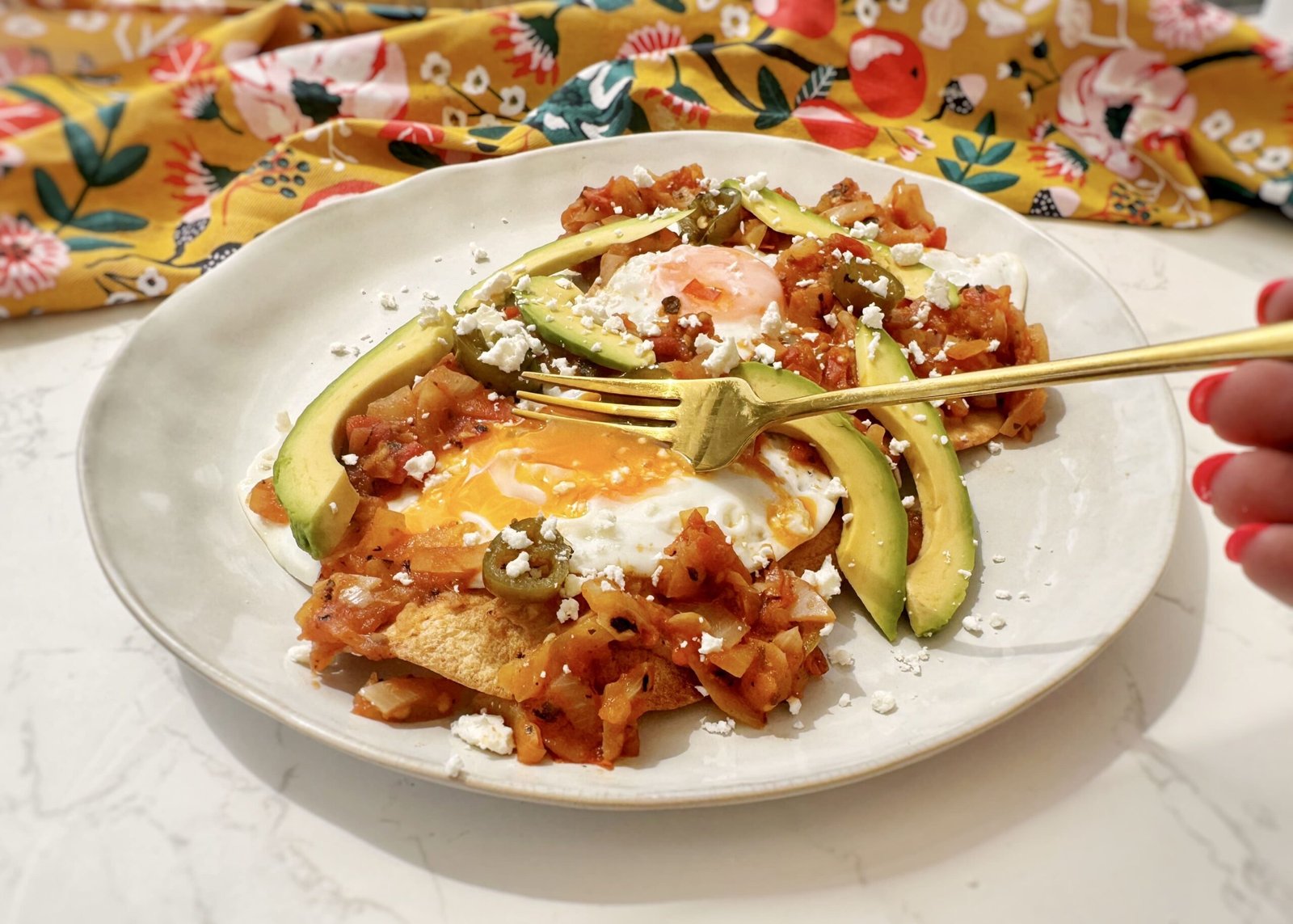 A plate of Huevos Rancheros with a crispy tortilla base, runny fried egg, refried beans, tomato salsa, avocado slices, and crumbled feta, garnished with coriander and lime wedges.