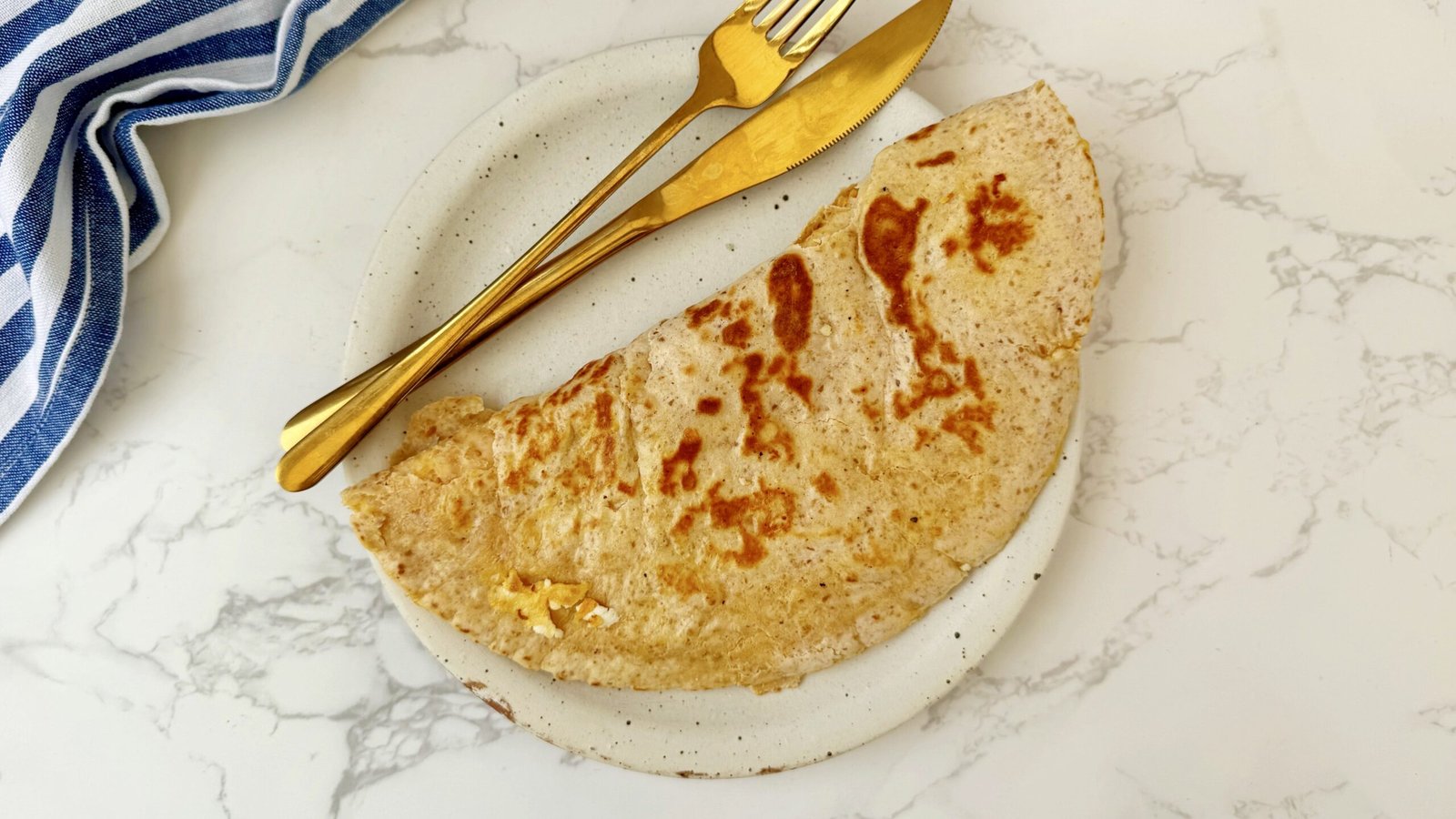 A folded tortilla omelette with a golden, crispy crust, showing melted feta, grated carrot, and fluffy egg layers. Served on a plate with fresh greens and lemon wedges.
