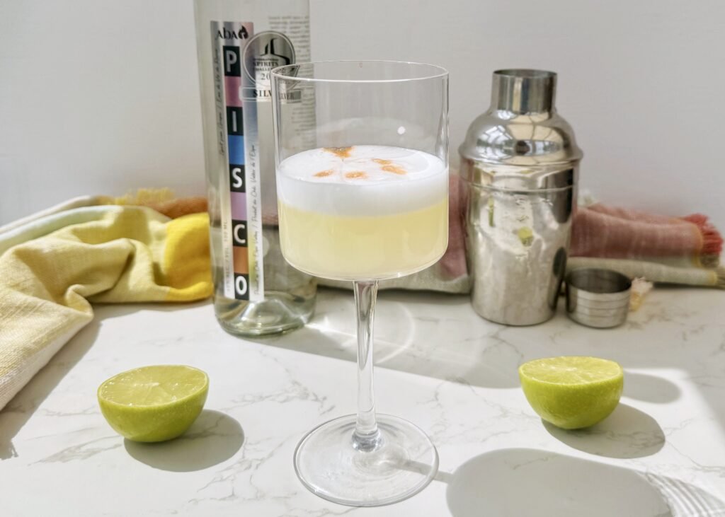 A frothy Pisco Sour cocktail in a chilled coupé glass, garnished with swirls of Angostura bitters on a creamy foam top, served with a lime wheel.