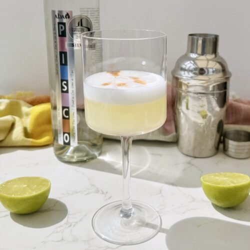 A frothy Pisco Sour cocktail in a chilled coupé glass, garnished with swirls of Angostura bitters on a creamy foam top, served with a lime wheel.