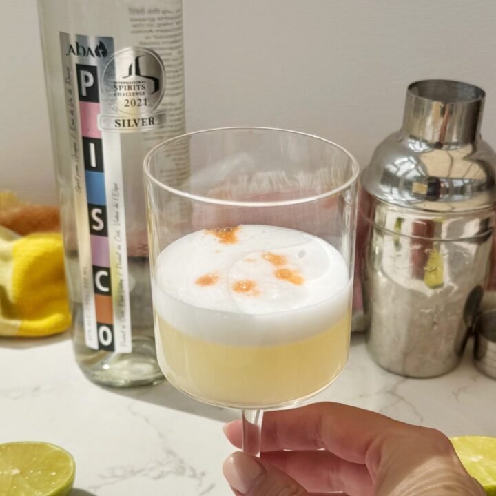 A frothy Pisco Sour cocktail in a chilled coupé glass, garnished with swirls of Angostura bitters on a creamy foam top, served with a lime wheel.