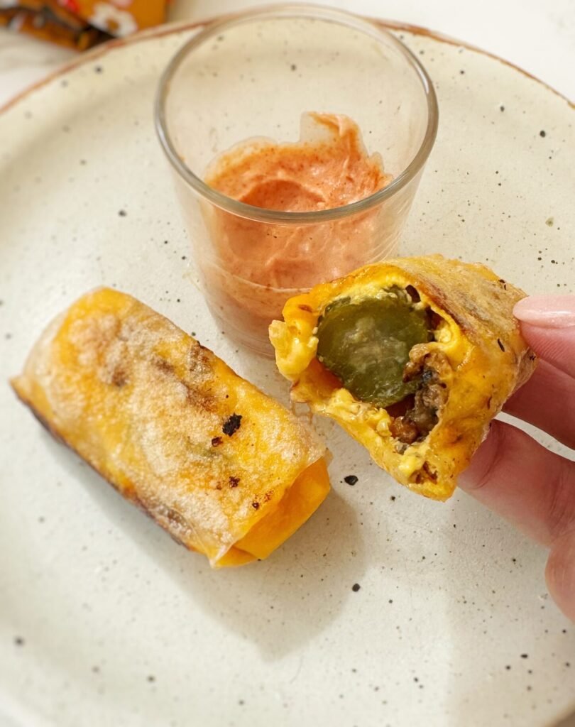 Crispy rice paper cheeseburger rolls filled with melted cheese, ground beef and pickles, served with dipping sauces on a platter.