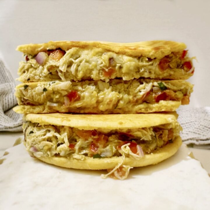Image of a toasted Colombian-style bun stuffed with mashed avocado, shredded chicken, tomato, and onion, topped with a crispy golden cheese crust, served on a rustic plate.