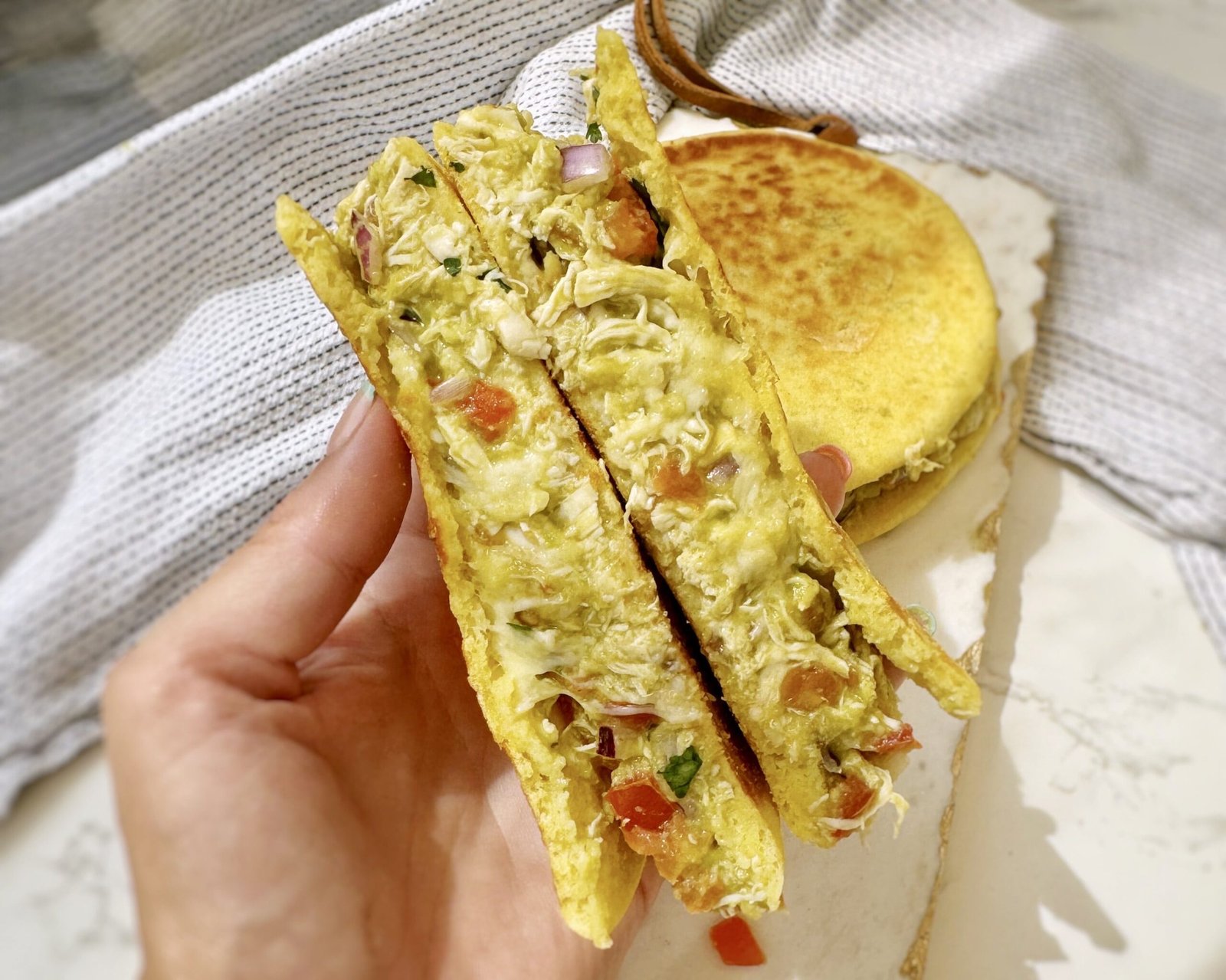 Image of a toasted Colombian-style bun stuffed with mashed avocado, shredded chicken, tomato, and onion, topped with a crispy golden cheese crust, served on a rustic plate.