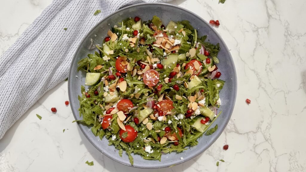 A fresh bowl of Arugula & Date Salad with arugula leaves, sliced Medjool dates, cherry tomatoes, feta, and toasted almonds drizzled with lemon-honey dressing.