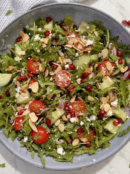 A fresh bowl of Arugula & Date Salad with arugula leaves, sliced Medjool dates, cherry tomatoes, feta, and toasted almonds drizzled with lemon-honey dressing.