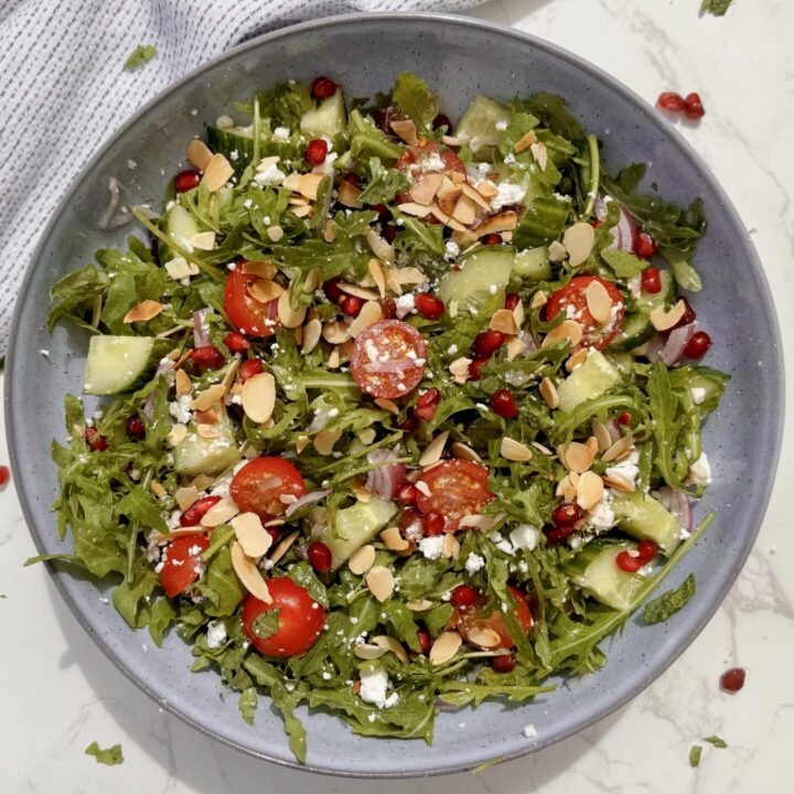 A fresh bowl of Arugula & Date Salad with arugula leaves, sliced Medjool dates, cherry tomatoes, feta, and toasted almonds drizzled with lemon-honey dressing.
