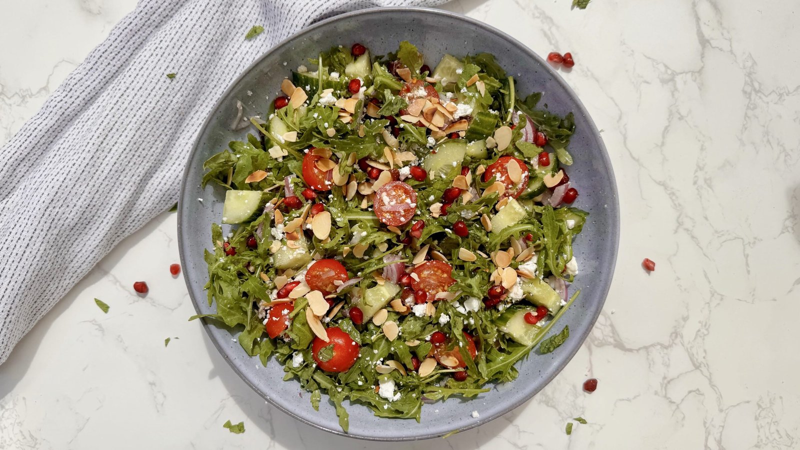A fresh bowl of Arugula & Date Salad with arugula leaves, sliced Medjool dates, cherry tomatoes, feta, and toasted almonds drizzled with lemon-honey dressing.