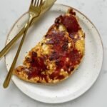 Close-up of crispy mozzarella and red pesto eggs in a frying pan, with golden cheese edges, vibrant red pesto base, and perfectly cooked sunny-side-up eggs. Preguntar a ChatGPT