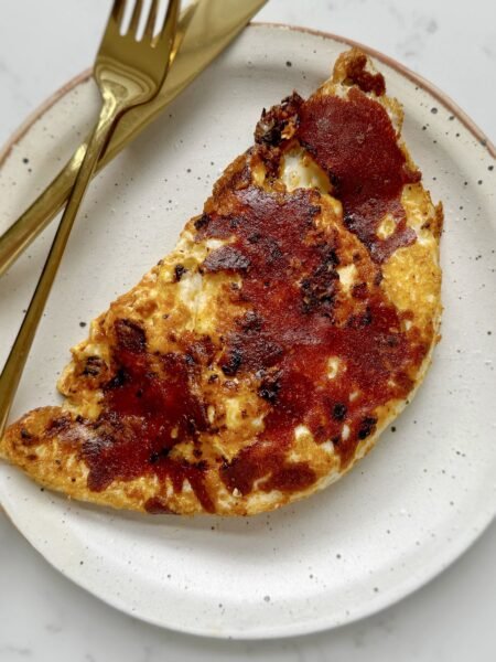Close-up of crispy mozzarella and red pesto eggs in a frying pan, with golden cheese edges, vibrant red pesto base, and perfectly cooked sunny-side-up eggs. Preguntar a ChatGPT