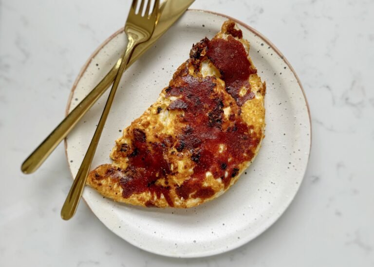Close-up of crispy mozzarella and red pesto eggs in a frying pan, with golden cheese edges, vibrant red pesto base, and perfectly cooked sunny-side-up eggs. Preguntar a ChatGPT