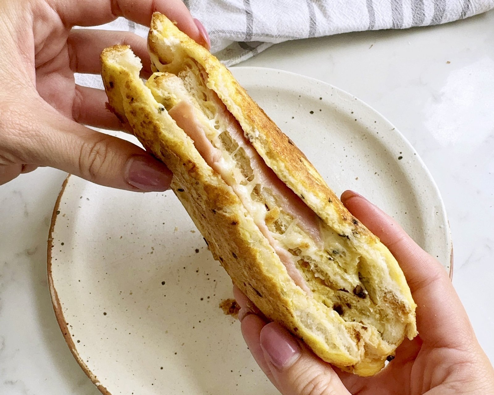Golden, crispy egg sandwich filled with fluffy scrambled eggs, melted cheese, and savoury ham, served hot on a white plate.