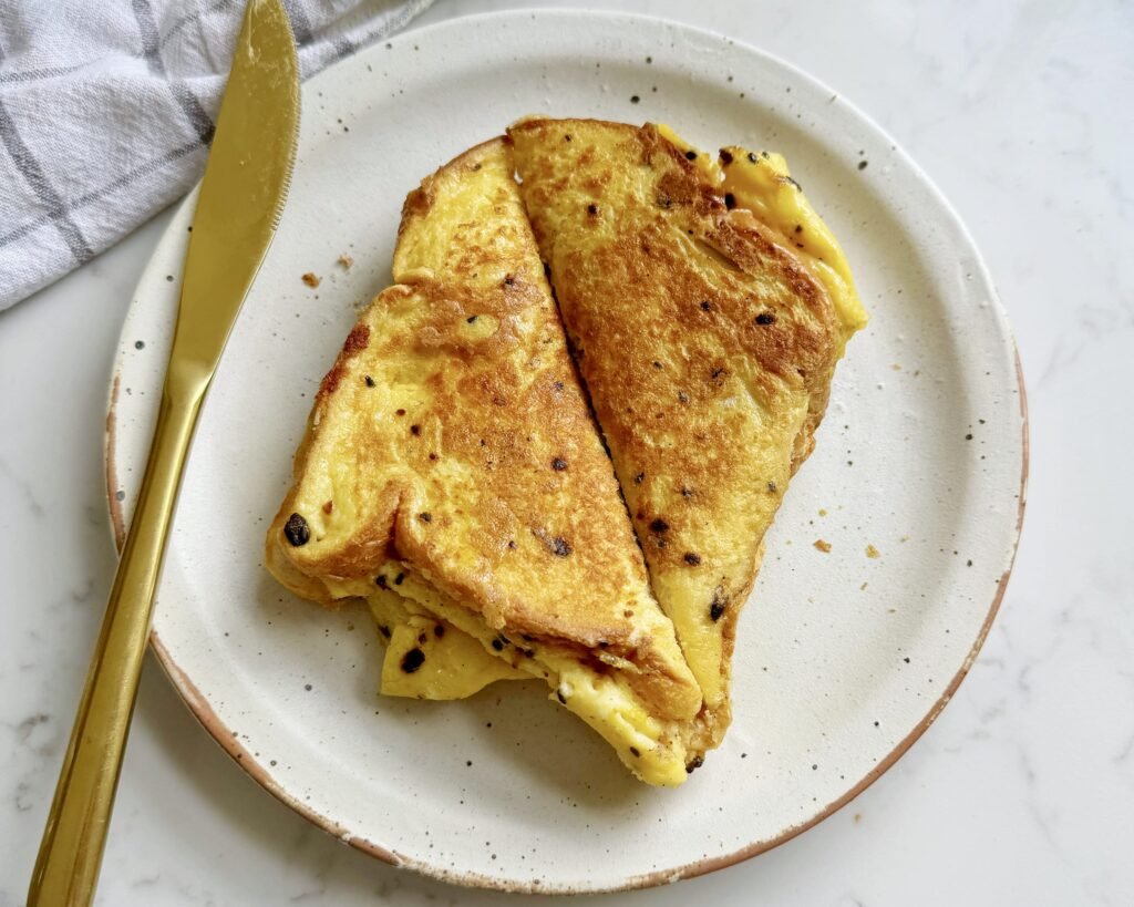 Golden, crispy egg sandwich filled with fluffy scrambled eggs, melted cheese, and savoury ham, served hot on a white plate.