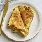 Golden, crispy egg sandwich filled with fluffy scrambled eggs, melted cheese, and savoury ham, served hot on a white plate.