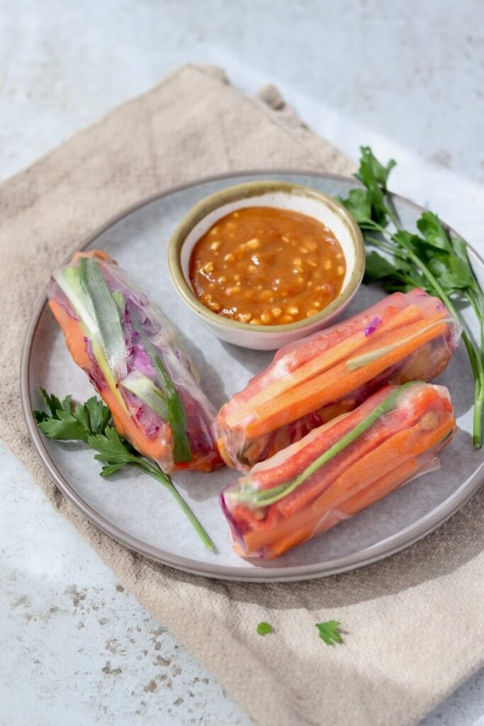 Three rice paper spring rolls filled with colourful julienned vegetables on a grey ceramic plate, served with a small bowl of chunky peanut satay dipping sauce and fresh parsley sprigs, styled on a beige linen cloth.