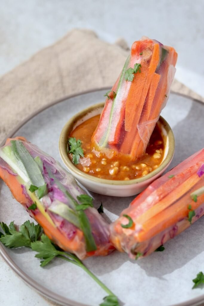 Three rice paper spring rolls filled with carrot, red pepper, cucumber, and red cabbage, arranged around a small bowl of peanut satay dipping sauce on a grey ceramic plate, with one roll dipped into the sauce and fresh parsley scattered around.