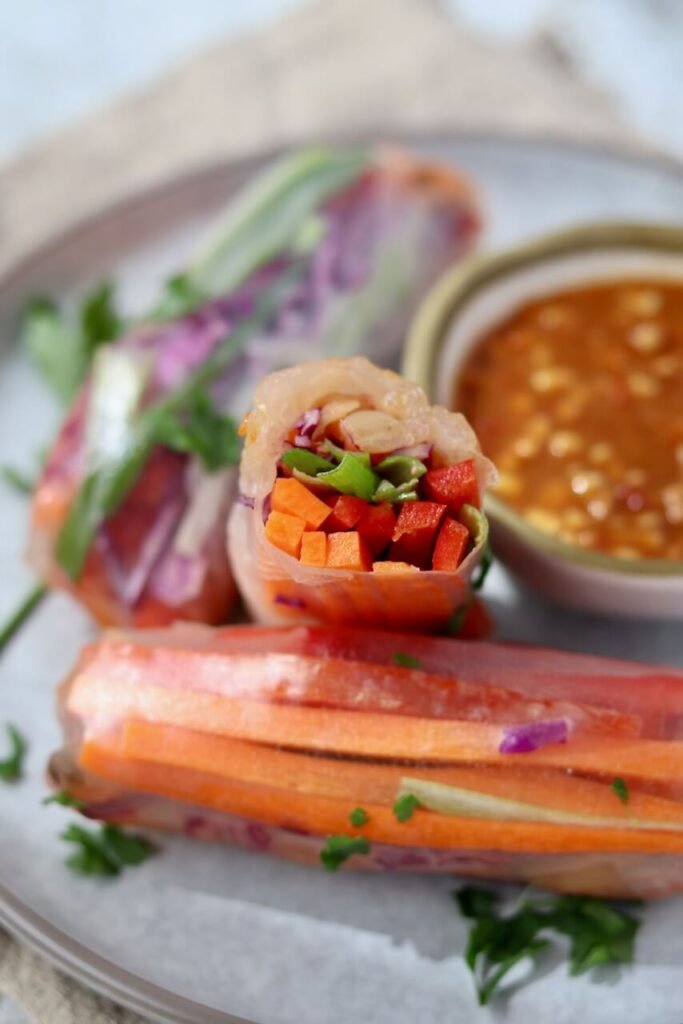 Close-up of a halved rice paper spring roll standing upright, revealing the colourful filling of diced carrot, red pepper, spring onion, and red cabbage, with whole rolls and a bowl of peanut satay sauce in the background.