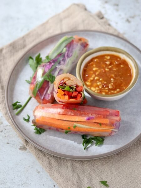 Close-up of a halved rice paper spring roll standing upright on a grey ceramic plate, revealing the colourful filling of diced carrot, red pepper, spring onion, and red cabbage, with whole rolls, fresh parsley, and a bowl of peanut satay dipping sauce arranged alongside on a beige linen cloth.