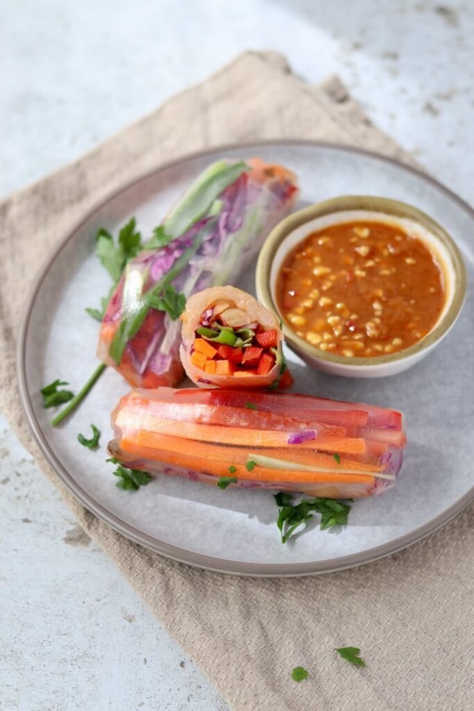 Close-up of a halved rice paper spring roll standing upright on a grey ceramic plate, revealing the colourful filling of diced carrot, red pepper, spring onion, and red cabbage, with whole rolls, fresh parsley, and a bowl of peanut satay dipping sauce arranged alongside on a beige linen cloth.