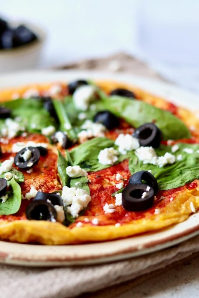 A close-up of a pizza topped with tomato sauce, fresh spinach leaves, black olives, and crumbled feta cheese on a plate.